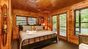 Een slaapkamer in een houten blokhut met een bed, nachtkastje, lamp en raam met jaloezieën.