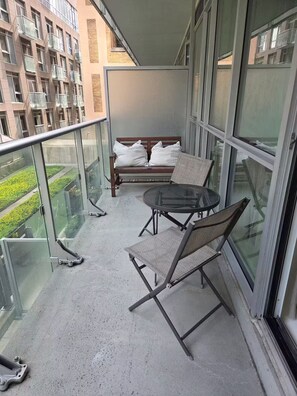 A balcony with a glass railing, a round table, two chairs, a sofa with cushions, and a view of a building with multiple windows. - Cozy 1-bedroom apartment with AC steps to attractions in downtown Toronto (Toronto)