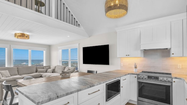 A modern kitchen with white cabinetry, a large island, a built-in oven, and a flat-screen TV. - Dune Life at OKI: Oceanfront with Pool (Oak Island)