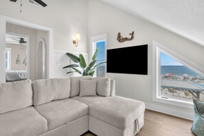Ein modernes Wohnzimmer mit einem Ecksofa, einem großen Fenster mit Blick auf den Strand, einem flachbildfernseher an der Wand und einer Zimmerpflanze.