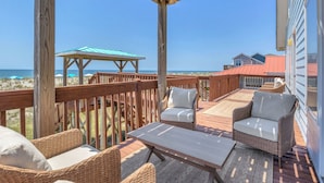 A wooden deck with wicker furniture and a table, overlooking the beach and ocean. - Dune Life at OKI: Oceanfront with Pool (Oak Island)