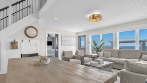 A spacious living area with a large wooden dining table, a sofa set, and a staircase with a railing. - Dune Life at OKI: Oceanfront with Pool (Oak Island)
