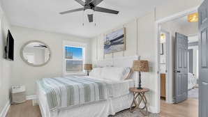 A bedroom with a bed, ceiling fan, bedside lamps, a round mirror, and a painting on the wall. - Dune Life at OKI: Oceanfront with Pool (Oak Island)
