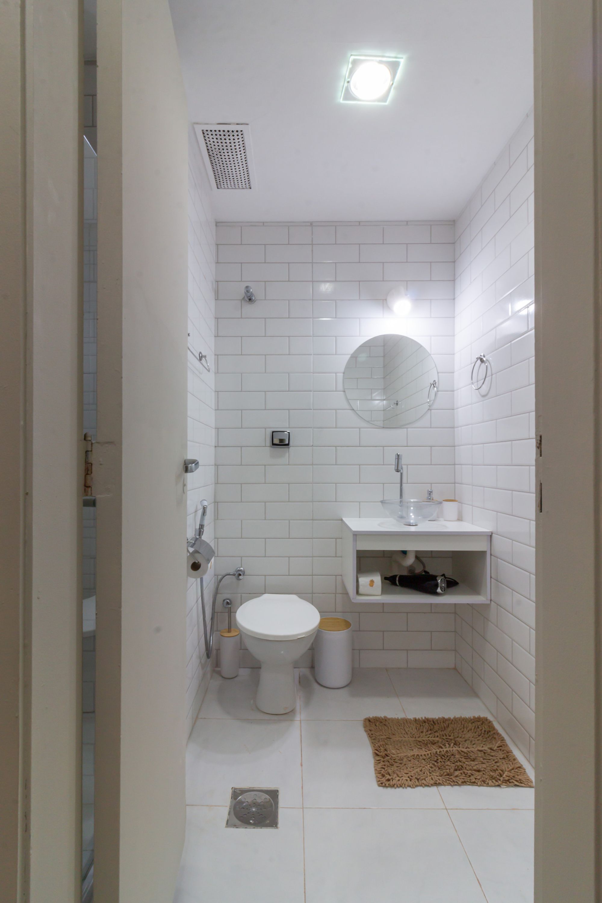 Un baño pequeño con azulejos blancos, que incluye inodoro, lavamanos, espejo y zona de ducha.