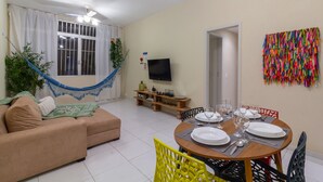 A living room with a sofa, a dining table set for four, a TV mounted on the wall, and a colorful wall decoration. - 8 min da Praia de Ipanema Bt705 (Rio de Janeiro)