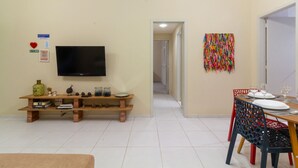 A living room with a TV, wooden entertainment unit, dining table with chairs, and a colorful wall hanging. - 8 min da Praia de Ipanema Bt705 (Rio de Janeiro)