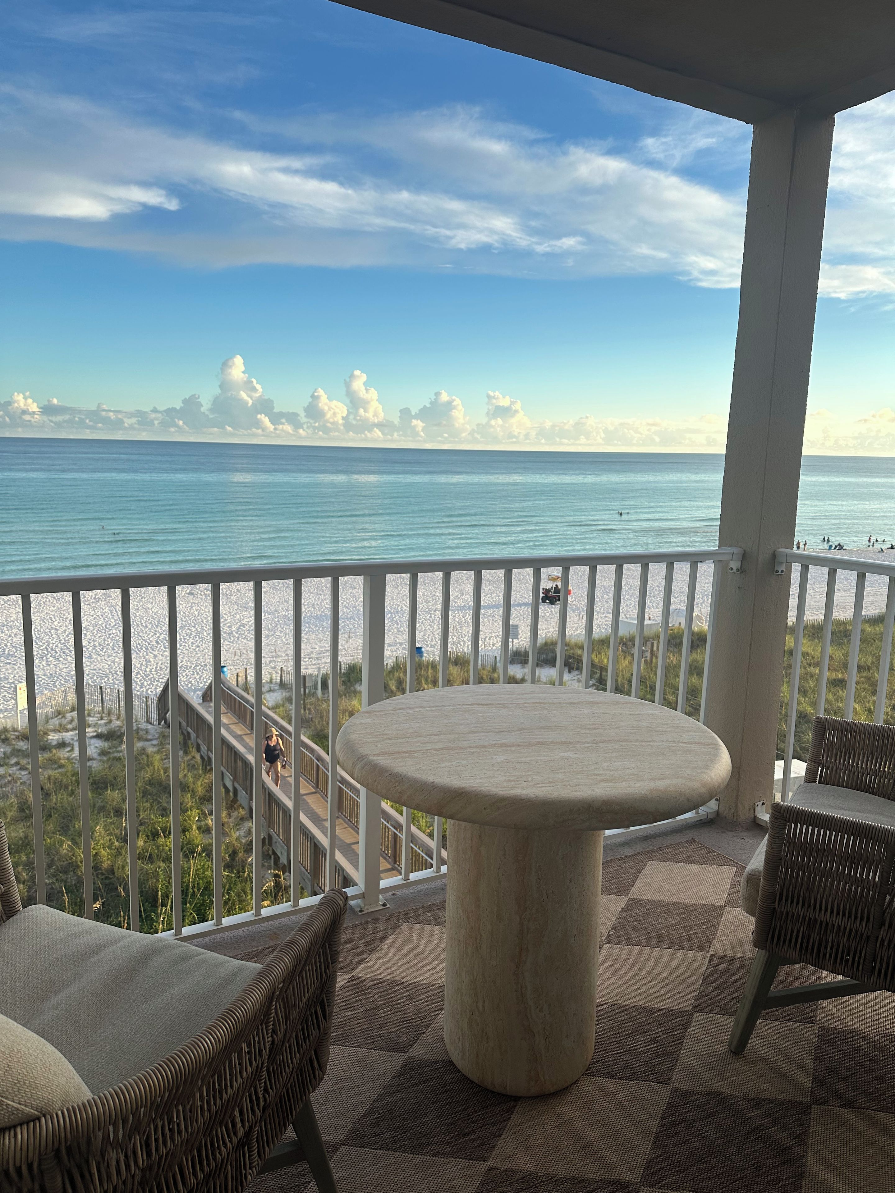 A balcony with a round table and wicker chairs, overlooking a beach and ocean.