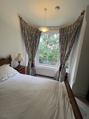 A bedroom with a large bed, a nightstand with a lamp, and a window with curtains. - Bluebell Suite at Elmwood House (Nottingham)