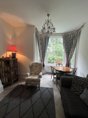 A living room with a sofa, armchair, table, chandelier, and a window with curtains. - Bluebell Suite at Elmwood House (Nottingham)