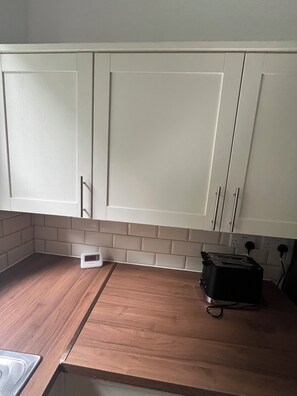 A kitchen with white cabinets, a wooden countertop, a digital clock, and a toaster. - Bluebell Suite at Elmwood House (Nottingham)