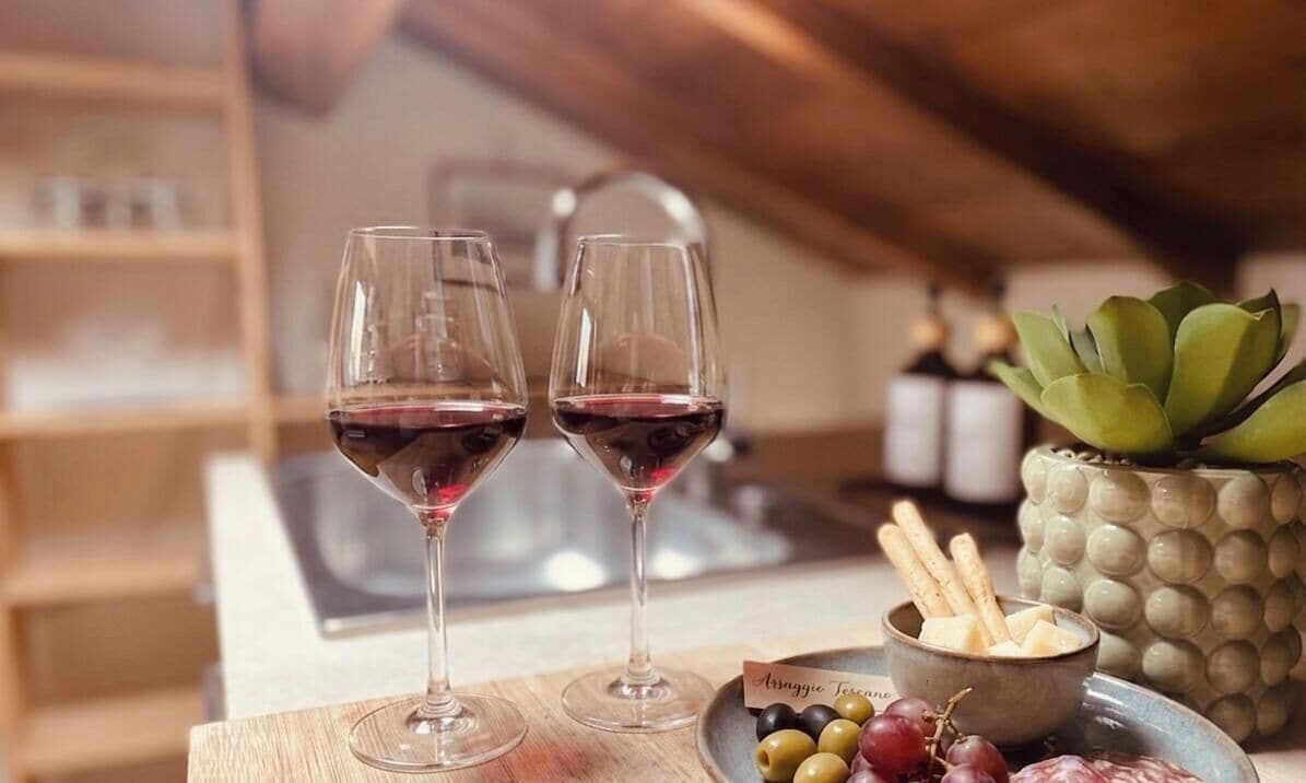 Two wine glasses on a wooden table with a bowl of olives and cheese, and a potted plant in the background.