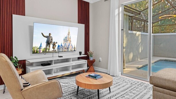 A modern living room with a flat-screen TV, a round coffee table, and a pool visible through glass doors. - 4BR Storey Lake Home Near Disney with Pool (Kissimmee)