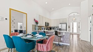 Una cocina moderna con armarios blancos, microondas y refrigerador. Hay un comedor con mesa de vidrio y sillas color turquesa.