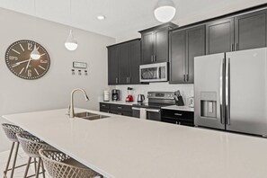 A modern kitchen with dark cabinets, stainless steel appliances, a large island with bar stools, and a wall clock. - 4BR Storey Lake Home Near Disney with Pool (Kissimmee)