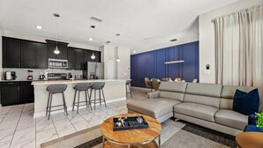 Et moderne køkken med mørke skabe, en central ø med barstole, et spiseområde med sofa og sofabord, samt en stue med hjørnesofa.