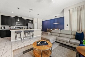A modern kitchen with dark cabinets, a central island with bar stools, a dining area with a sofa and coffee table, and a living room with a sectional sofa. - 4BR Storey Lake Home Near Disney with Pool (Kissimmee)