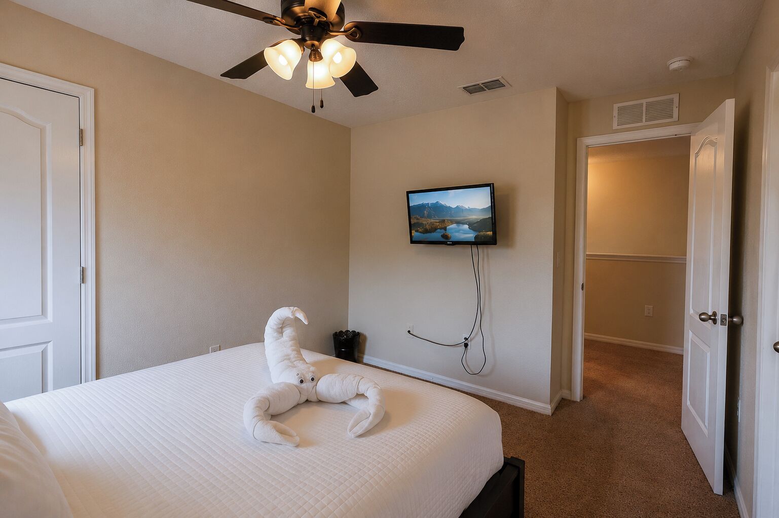 A bedroom with a bed, a ceiling fan, a mounted TV, and a door leading to another room.