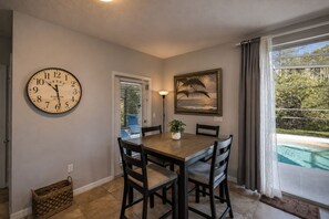 A dining area with a wooden table, four chairs, a wall clock, a framed picture, and a view of a swimming pool. - Meadow Manor - 10 Mins to Disney! Pool/spa (Kissimmee)