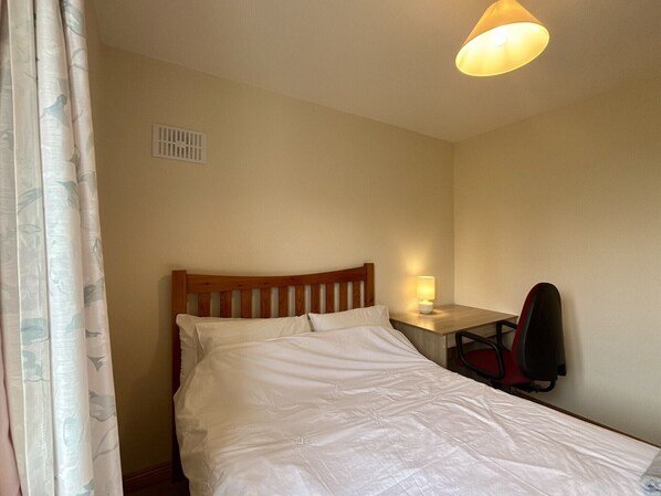 A single bed with a wooden headboard, a bedside table with a lamp, a chair, and a window with curtains.
