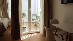Ein Zimmer mit einem Bett, einem Tisch und einem Stuhl. Eine Schiebetür öffnet sich zu einem Außenbereich mit einem Stuhl und einer Pflanze.