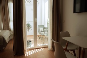 Ein Zimmer mit einem Bett, einem Tisch und einem Stuhl. Eine Schiebetür öffnet sich zu einem Außenbereich mit einem Stuhl und einer Pflanze.