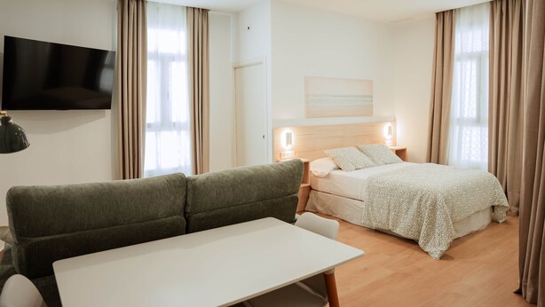 A modern hotel room with a sofa, a bed, a flat-screen TV, and a painting on the wall. - Olea Suites Conil · 4 (Conil de la Frontera)