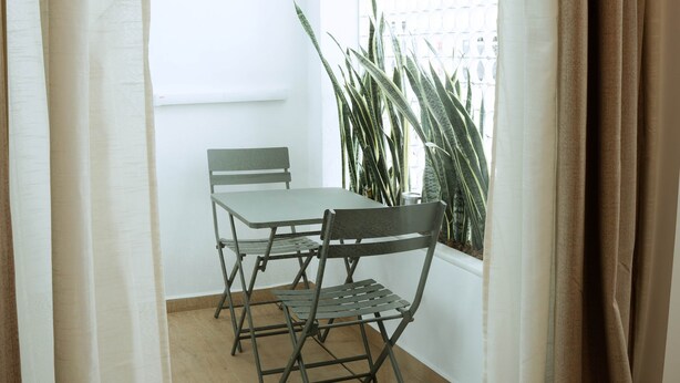A room with a table and chairs, a window with curtains, and a plant. - Olea Suites Conil · 5 (Conil de la Frontera)