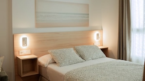 A hotel room with a bed, two bedside tables, a wall-mounted light, and a painting above the bed. - Olea Suites Conil · 2 (Conil de la Frontera)