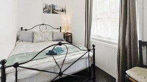 A neatly arranged bedroom with a metal bed frame, a bedside table with a lamp, a window with curtains, and a wall-mounted picture. - Park View En-Suite I Egham station (1 miunte walk) (Egham)