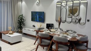 A modern dining area with a wooden table, grey upholstered chairs, and a large flat-screen TV mounted on the wall. - 2 BR SLEEPS 6 PAX INDOOR POOL, GYM AND BEAUTIFUL NAIROBI CITY VIEWS (Nairobi)