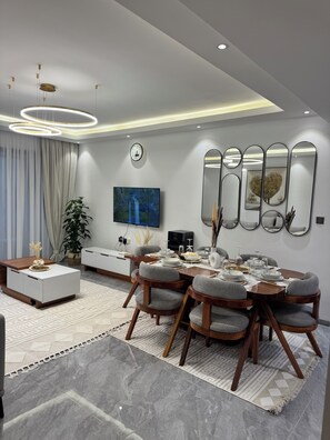 Uma área de jantar moderna com mesa de madeira, cadeiras estofadas cinzentas e uma grande televisão de tela plana na parede.