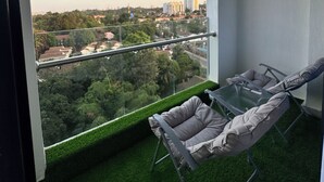 A balcony with a glass table and two chairs, overlooking a cityscape with buildings and greenery. - 2 BR SLEEPS 6 PAX INDOOR POOL, GYM AND BEAUTIFUL NAIROBI CITY VIEWS (Nairobi)