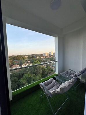 Uma varanda com mesa de vidro e duas cadeiras, com vista para um horizonte urbano com edifícios e áreas verdes.