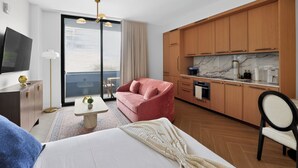 Ett modernt hotellrum med en säng, en röd soffa, ett litet bord, en TV, ett köksutrymme med träskåp och ett stort fönster.