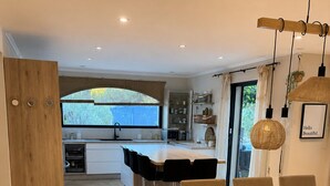 A modern kitchen with a large island, a dining area with a white table and chairs, and a view through the window.