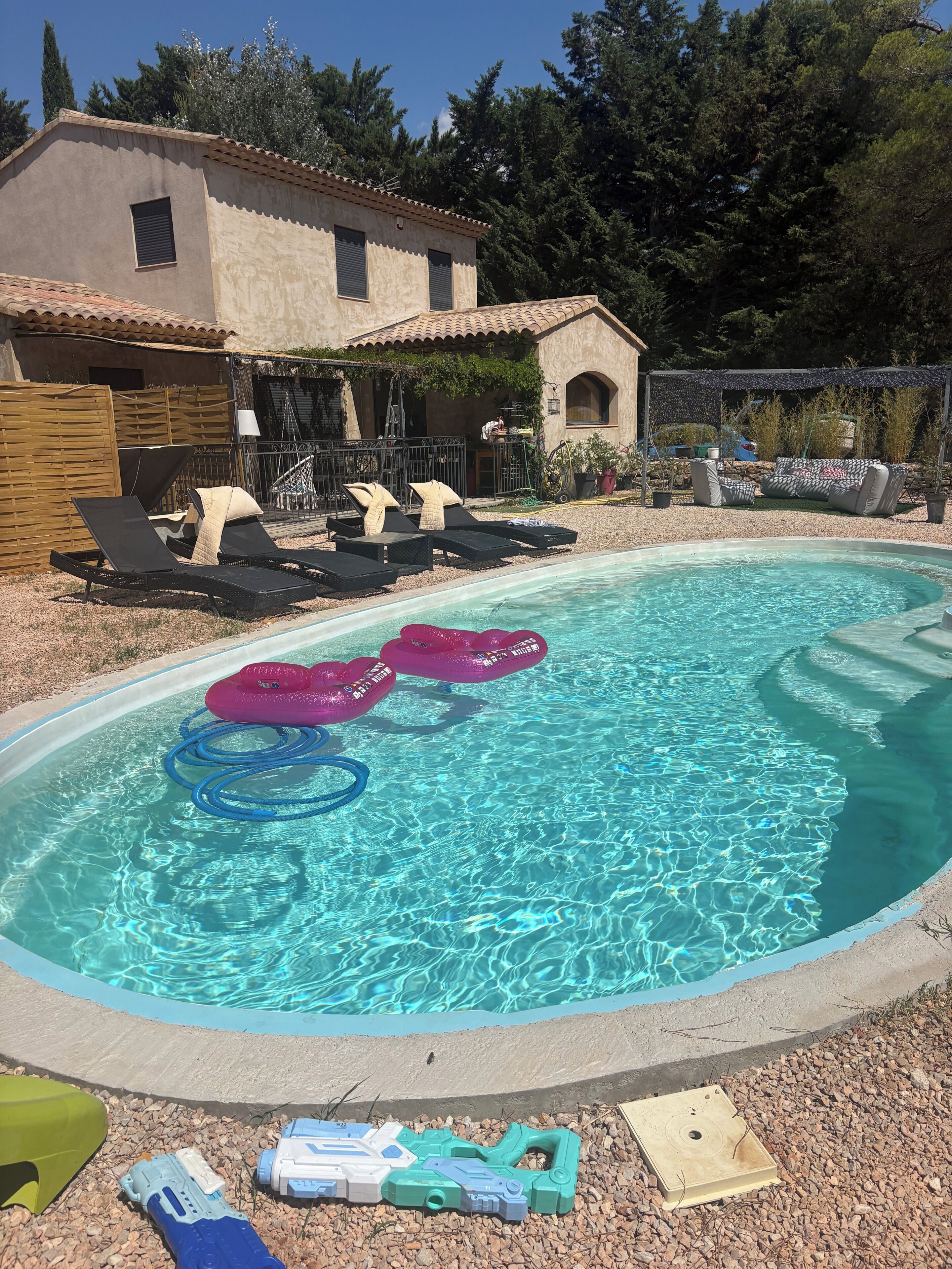 Une piscine extérieure dotée de chaises longues et de bouées gonflables, entourée d’une terrasse et avec un bâtiment visible en arrière-plan.
