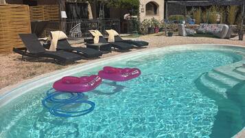 Une piscine extérieure dotée de chaises longues et de bouées gonflables, entourée d’une terrasse et avec un bâtiment visible en arrière-plan.