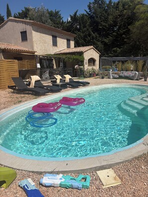 Une piscine extérieure dotée de chaises longues et de bouées gonflables, entourée d’une terrasse et avec un bâtiment visible en arrière-plan.