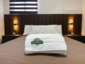 Una cama bien hecha con una manta blanca y una toalla con el texto 'La Antigua Estacion', cabecera de madera y luces fijadas a la pared.