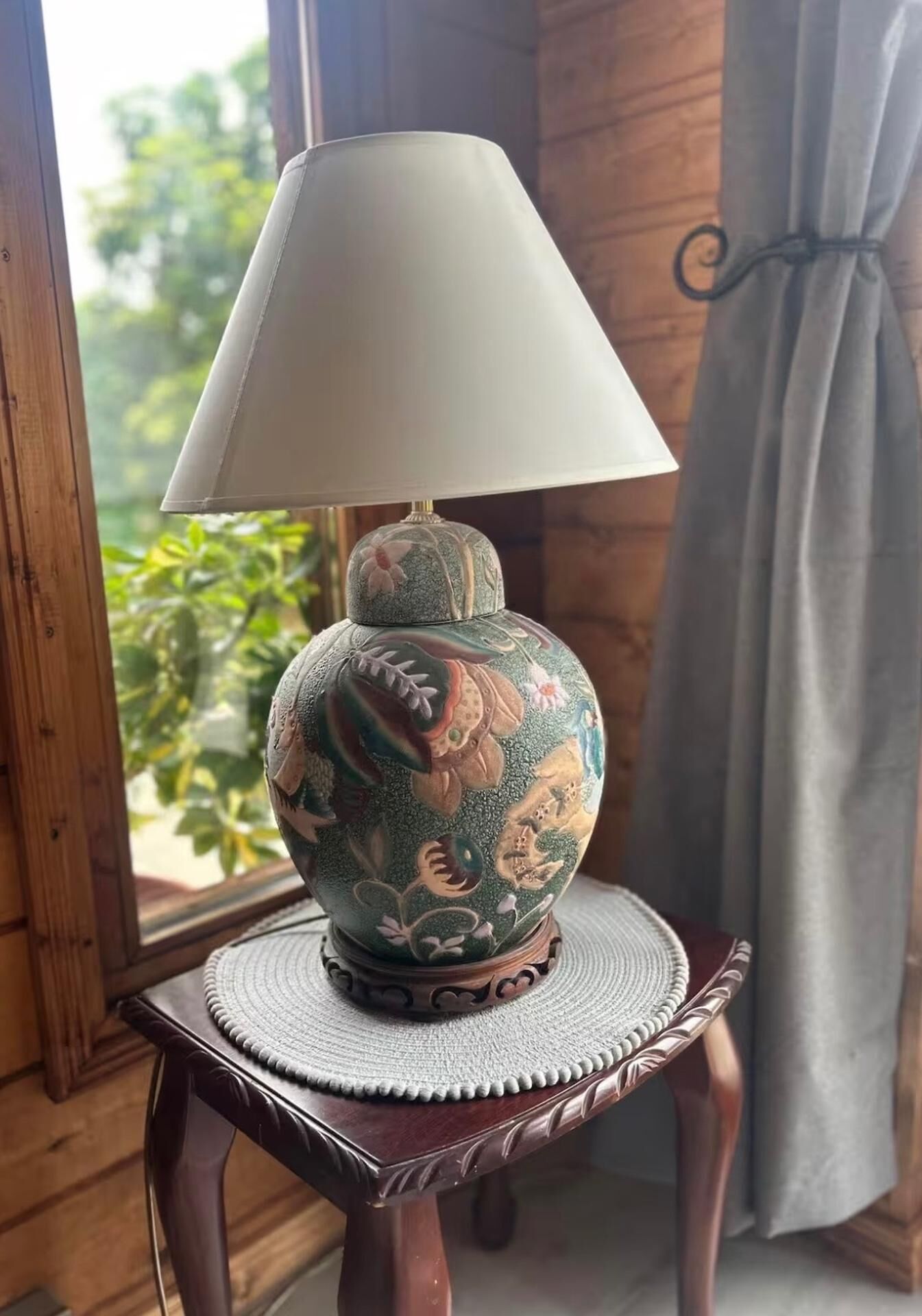Une lampe de table dotée d’un pied décoratif, posée sur une table ronde, près d’une fenêtre donnant sur un paysage verdoyant.