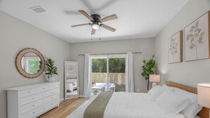 Een slaapkamer met een bed, een plafondventilator, een dressoir, een raam met gordijnen en uitzicht op de omgeving.