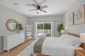 Ein Schlafzimmer mit Bett, Deckenventilator, Kommode, Fenster mit Vorhängen und Blick ins Freie.