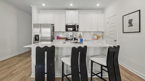 A modern kitchen with white cabinets, a stainless steel refrigerator, a microwave, and a large island with bar stools. - Grant & Groove: Where timeless design meets the rhythm of ATL. (Atlanta)