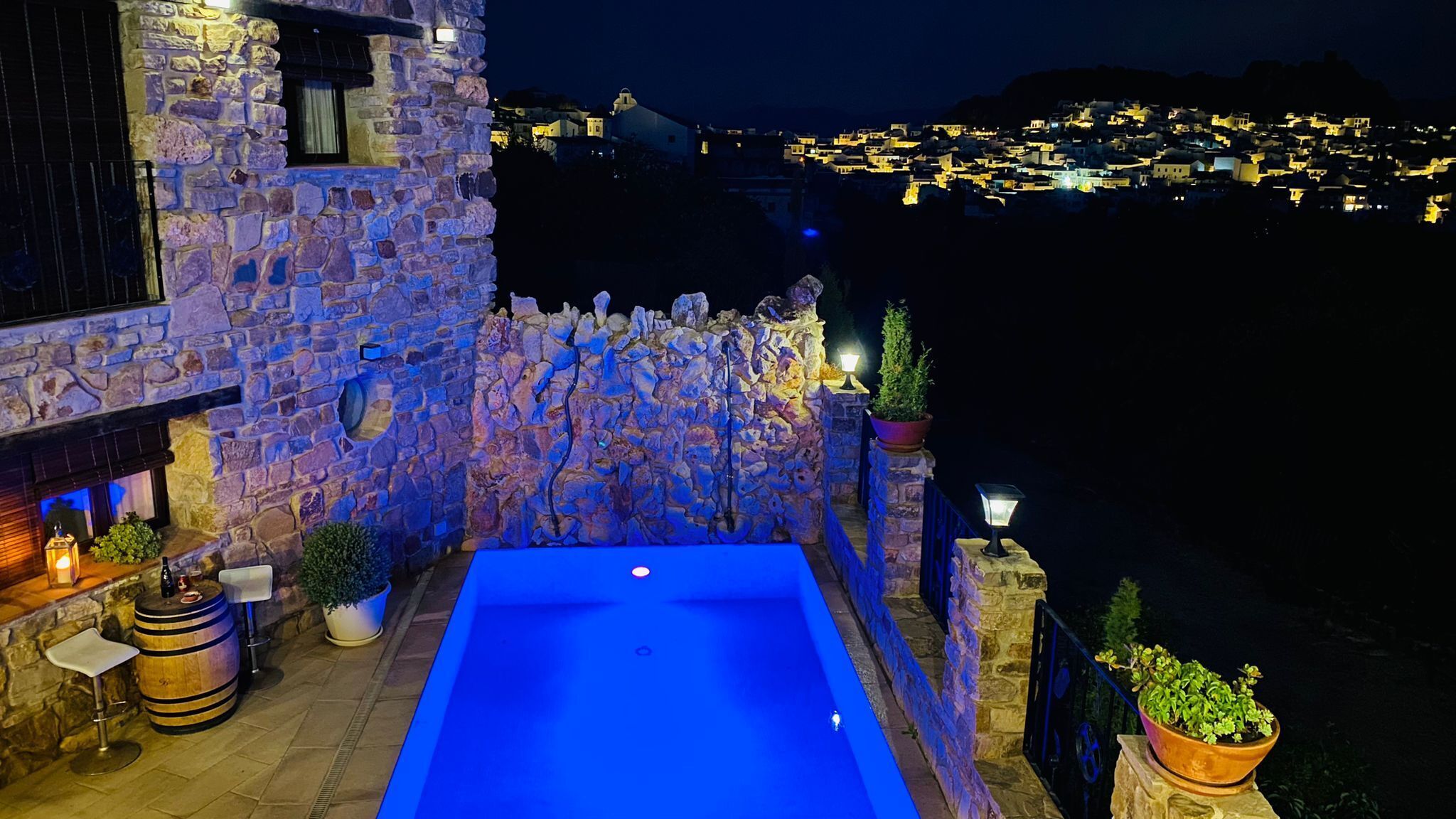 Edificio de piedra con piscina, plantas en macetas y vistas a un pueblo por la noche.