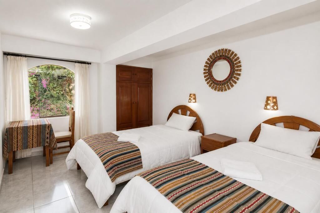 A hotel room with two beds, a wooden wardrobe, a small table with a patterned tablecloth, and a window with curtains.