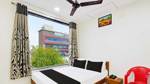 A hotel room with a bed, a desk, a chair, a ceiling fan, and a window with curtains. - HOTEL KALYAN (Rajkot)