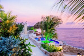 Zona de piscina con pared de piedra, palmeras y vista al mar al atardecer.