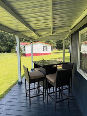 Un patio techado con una mesa y sillas, vista de una casa al fondo y un jardín con césped.