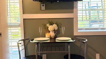 Un coin repas avec une table dressée pour deux personnes, un téléviseur à écran plat fixé au mur au-dessus, et une fenêtre équipée de stores.
