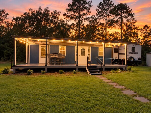 A mobile home with a deck, outdoor seating, and a Winnebago RV parked nearby.